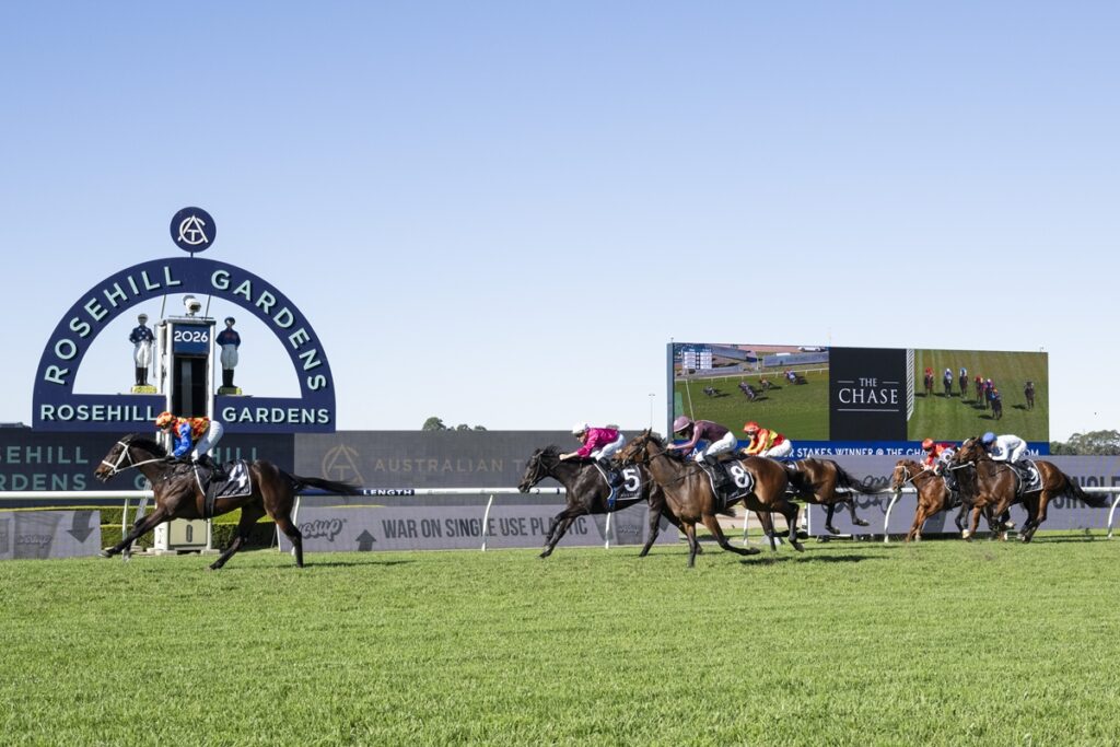 Horses and jockeys race down the track at Rosehill Gardens, with a large circular sign and video screen in the background on a sunny day.