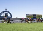 Horses and jockeys race down the track at Rosehill Gardens, with a large circular sign and video screen in the background on a sunny day.