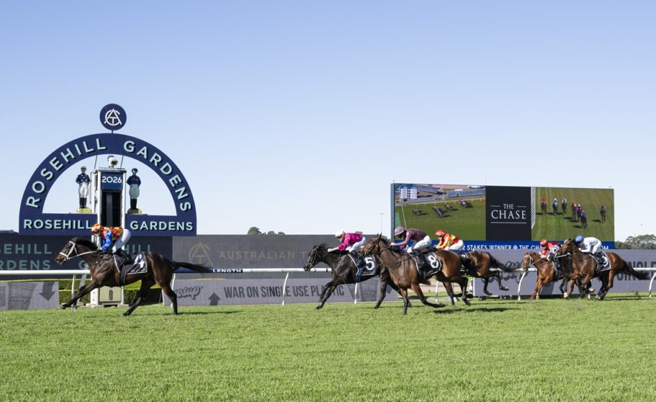 Horses and jockeys race down the track at Rosehill Gardens, with a large circular sign and video screen in the background on a sunny day.