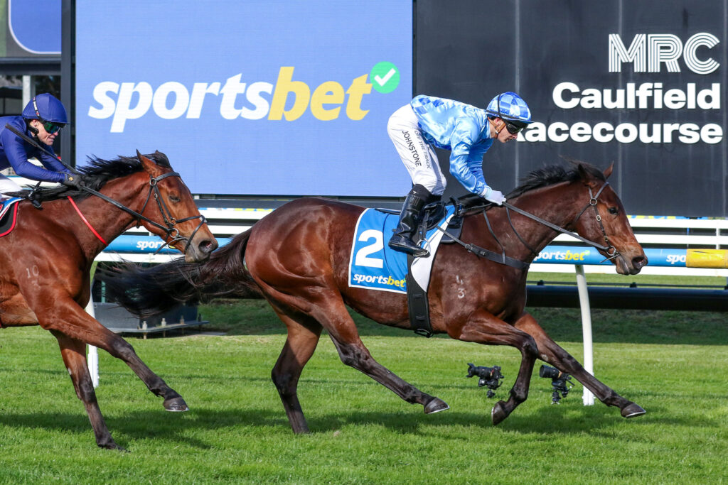 Two jockeys race brown horses on a grassy track, blue and white silks, number 2 saddle cloth, with a large 'sportsbet' sign in the background.