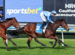 Two jockeys race brown horses on a grassy track, blue and white silks, number 2 saddle cloth, with a large 'sportsbet' sign in the background.