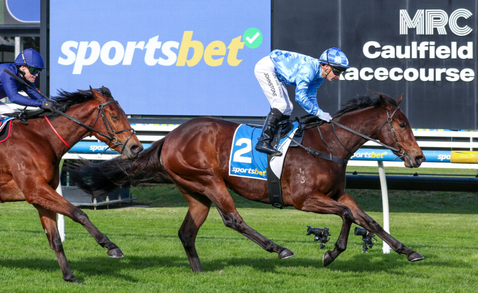Two jockeys race brown horses on a grassy track, blue and white silks, number 2 saddle cloth, with a large 'sportsbet' sign in the background.
