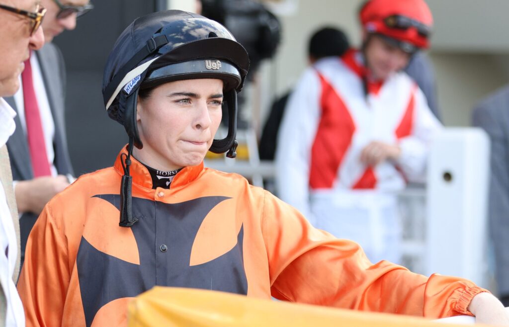 Close-up of a jockey in orange racing silks and a black helmet, looking ahead with lips pressed together at the trackside.