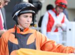 Close-up of a jockey in orange racing silks and a black helmet, looking ahead with lips pressed together at the trackside.