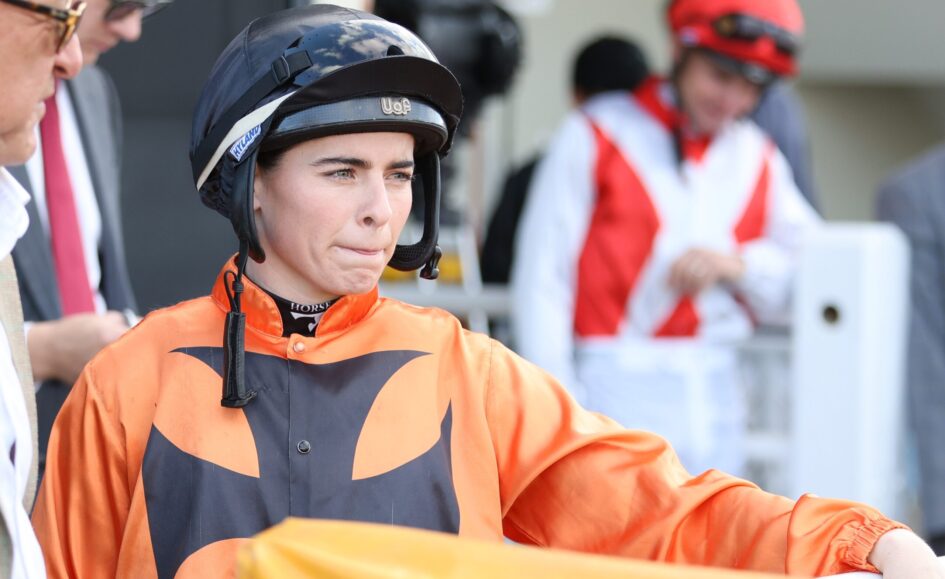 Close-up of a jockey in orange racing silks and a black helmet, looking ahead with lips pressed together at the trackside.