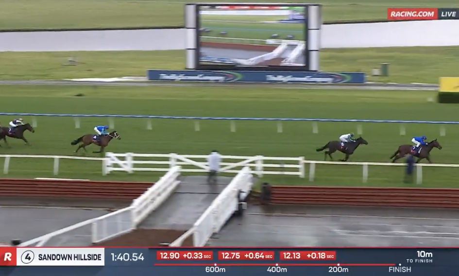 Horse race on turf at Sandown Hillside, blue-sleeved jockey leads a pack toward the finish with a large video screen in the background.