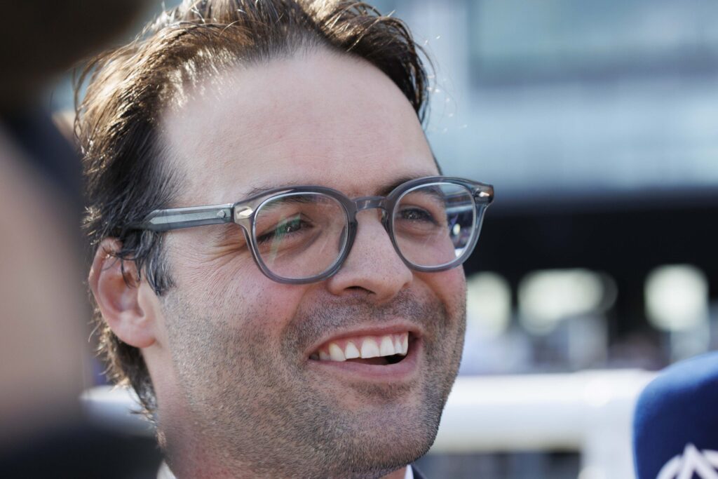 Close-up of a man smiling, wearing round glasses, cropped hair, outdoors in bright light.