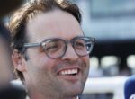 Close-up of a man smiling, wearing round glasses, cropped hair, outdoors in bright light.