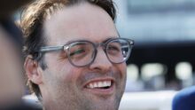 Close-up of a man smiling, wearing round glasses, cropped hair, outdoors in bright light.