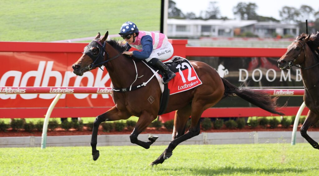Jockey in pink and white silks riding a brown racehorse during a race along a red railing.