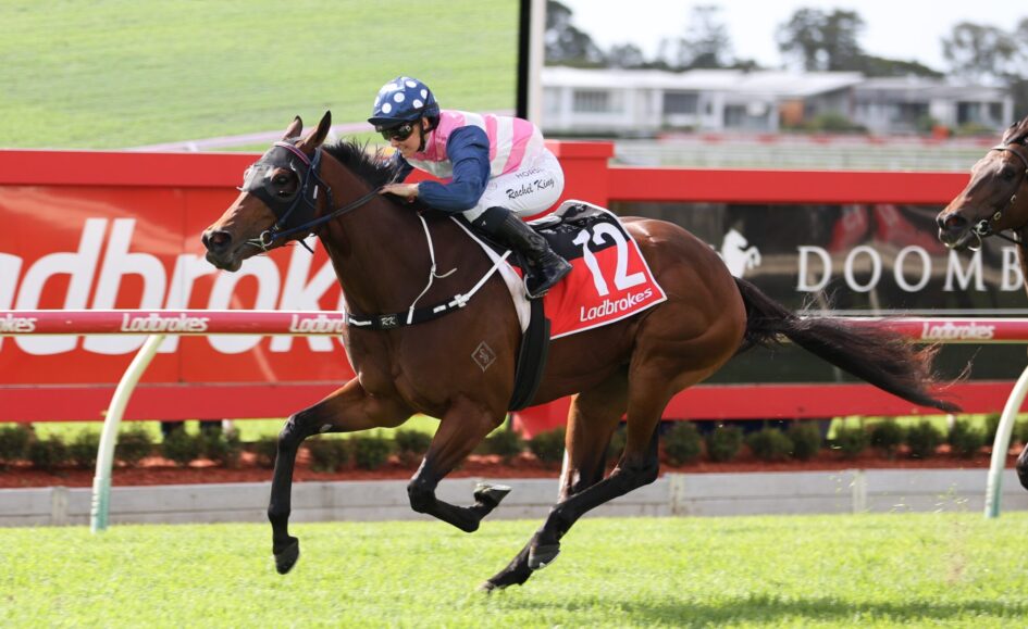 Jockey in pink and white silks riding a brown racehorse during a race along a red railing.