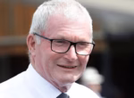 Smiling older man with white hair and black glasses, wearing a white shirt and tie outdoors.
