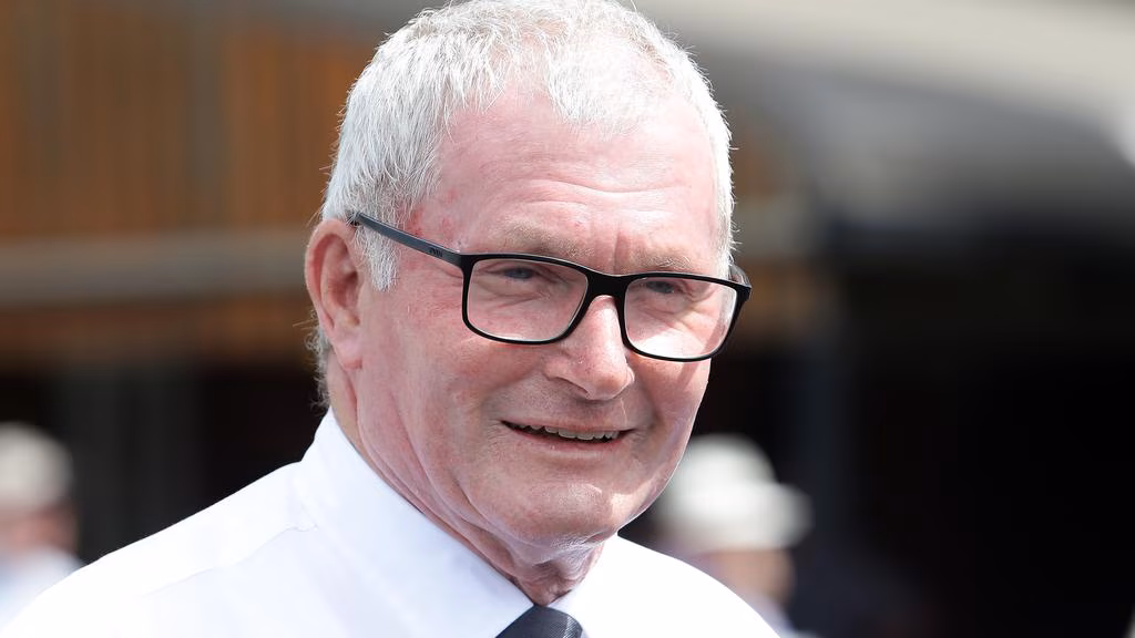 Smiling older man with white hair and black glasses, wearing a white shirt and tie outdoors.