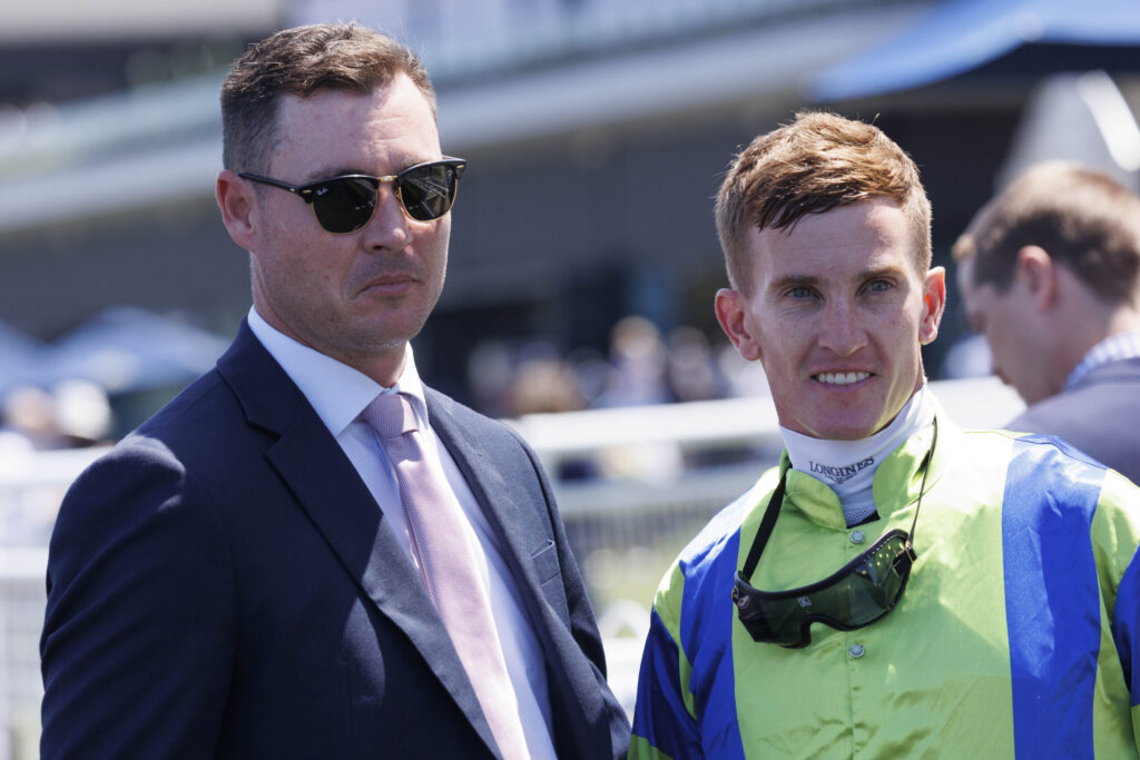 Man in a navy suit and pink tie wearing sunglasses stands beside a smiling worker in a bright green safety jacket and helmet, outdoors at an event.
