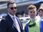 Man in a navy suit and pink tie wearing sunglasses stands beside a smiling worker in a bright green safety jacket and helmet, outdoors at an event.