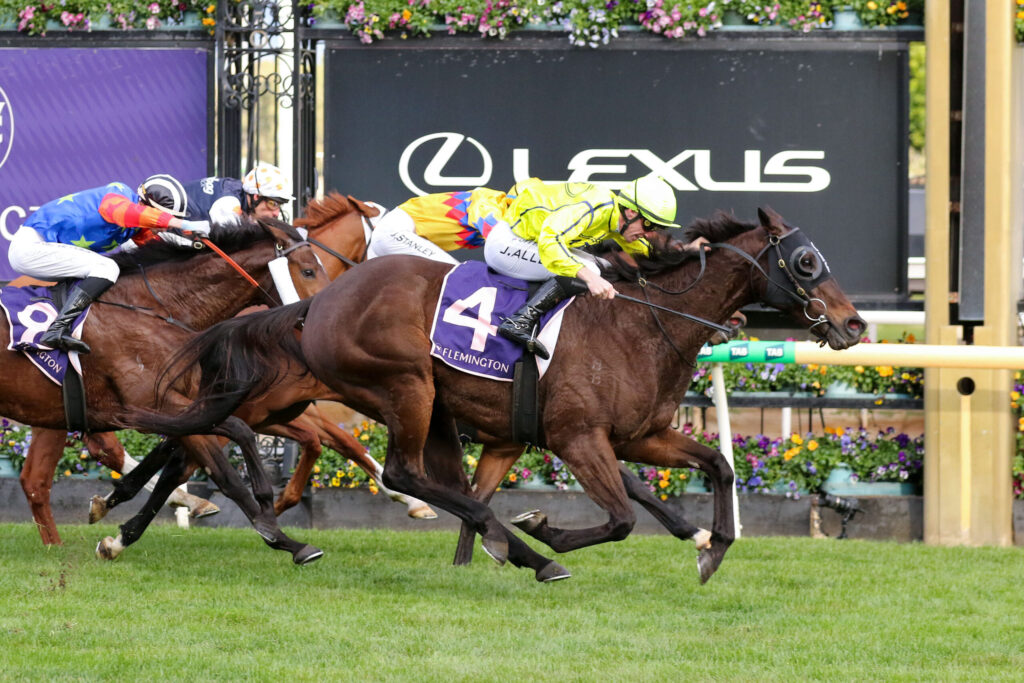 Horses and jockeys racing side by side on a grassy track with colorful silks and a dark sponsor banner in the background.