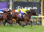 Horses and jockeys racing side by side on a grassy track with colorful silks and a dark sponsor banner in the background.