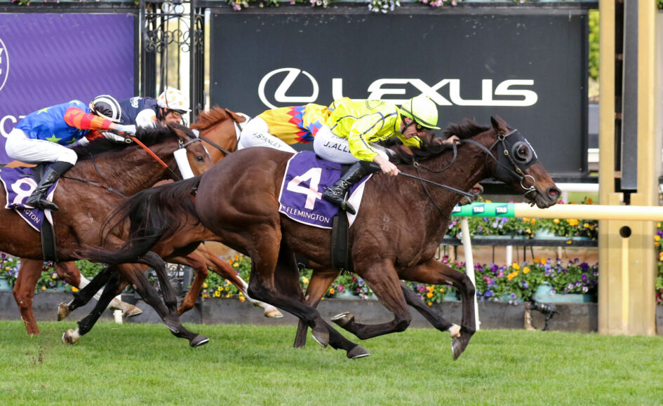 Horses and jockeys racing side by side on a grassy track with colorful silks and a dark sponsor banner in the background.