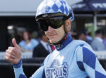 Jockey in a blue racing suit and checkered helmet gives a thumbs-up gesture at a race event.