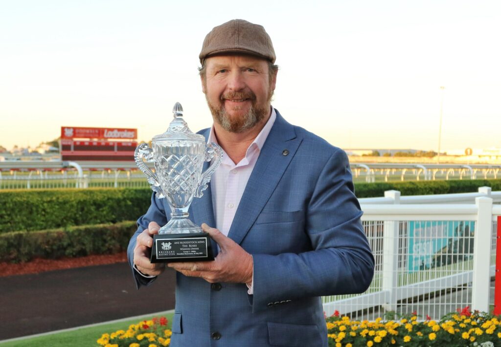 Man in a blue suit holding a crystal trophy outdoors at a racecourse.