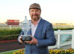 Man in a blue suit holding a crystal trophy outdoors at a racecourse.