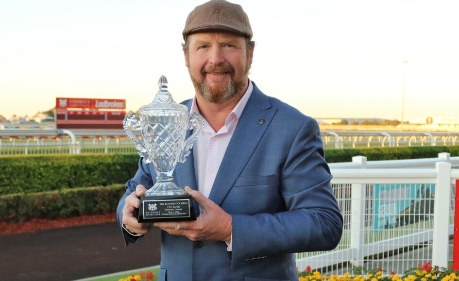 Man in a blue suit holding a crystal trophy outdoors at a racecourse.