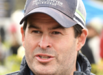 Close-up of a man wearing a gray cap and jacket, speaking outdoors.