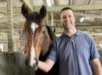 Man in a blue polo standing beside a brown horse, smiling, with his arm around the horse in a stable setting.