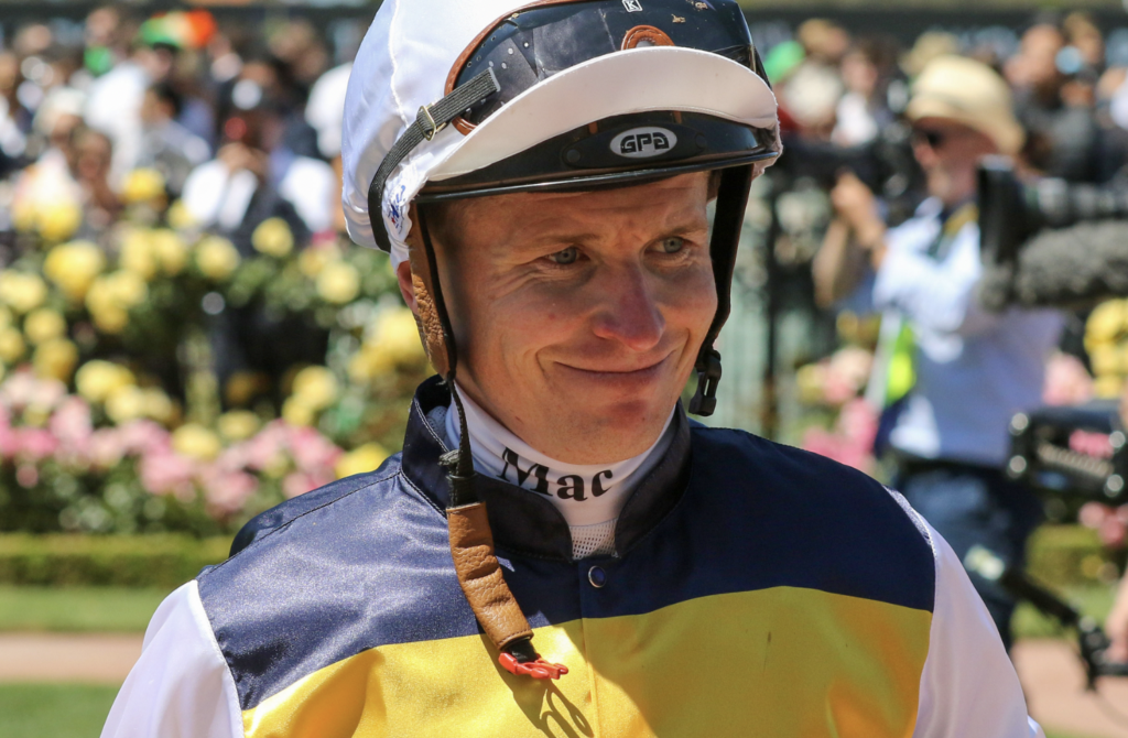 Jockey in a yellow and navy racing outfit smiles with helmet on, crowd behind at a racecourse