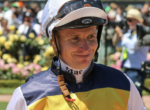 Jockey in a yellow and navy racing outfit smiles with helmet on, crowd behind at a racecourse
