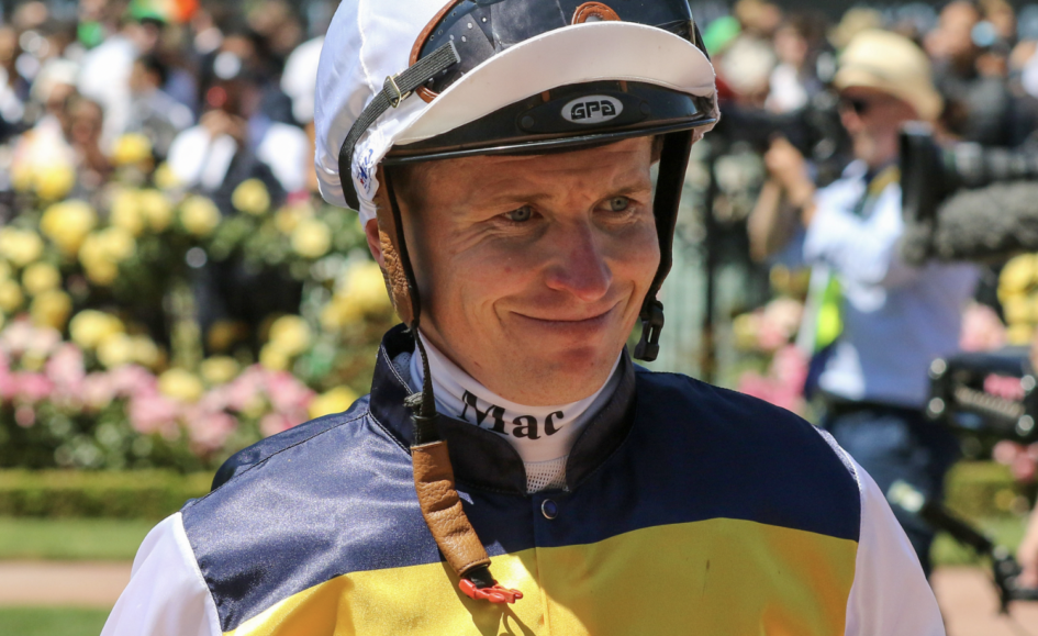 Jockey in a yellow and navy racing outfit smiles with helmet on, crowd behind at a racecourse