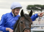 Man in a blue riding jacket and white helmet on a dark brown horse, smiling as he points to the right.