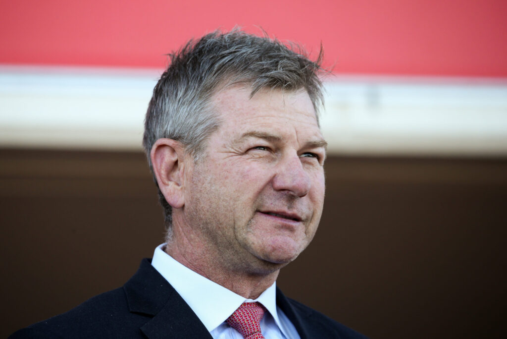Close-up of a middle-aged man in a dark suit and red tie, smiling, with a red wall background behind him.