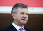 Close-up of a middle-aged man in a dark suit and red tie, smiling, with a red wall background behind him.