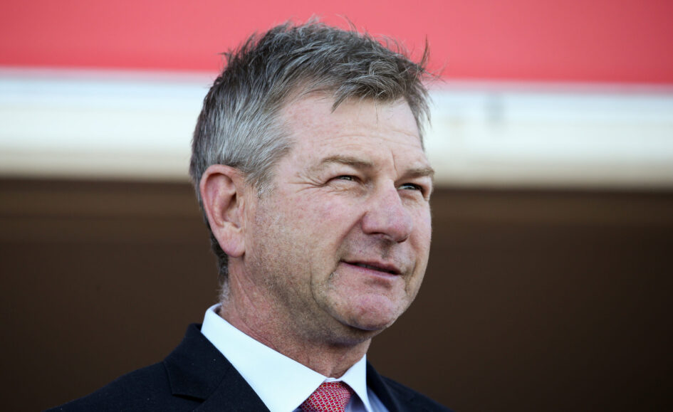 Close-up of a middle-aged man in a dark suit and red tie, smiling, with a red wall background behind him.