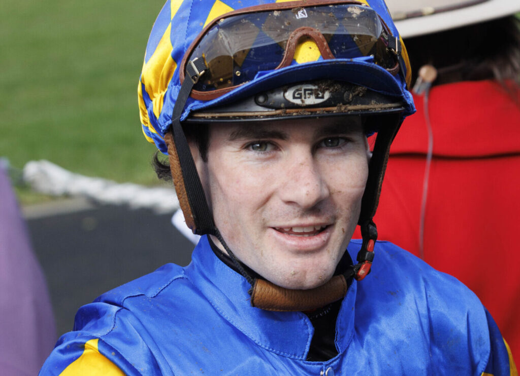 Close-up of a jockey wearing a blue and yellow helmet with goggles, facing the camera.