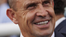 Close-up portrait of a smiling man in a dark suit and white shirt, showing teeth and laugh lines on his face.