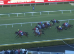 Horse race on a green turf track with jockeys in colorful silks chasing the lead near the inside rail; digital ad boards in background.