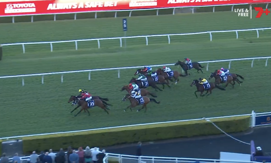 Horse race on a green turf track with jockeys in colorful silks chasing the lead near the inside rail; digital ad boards in background.