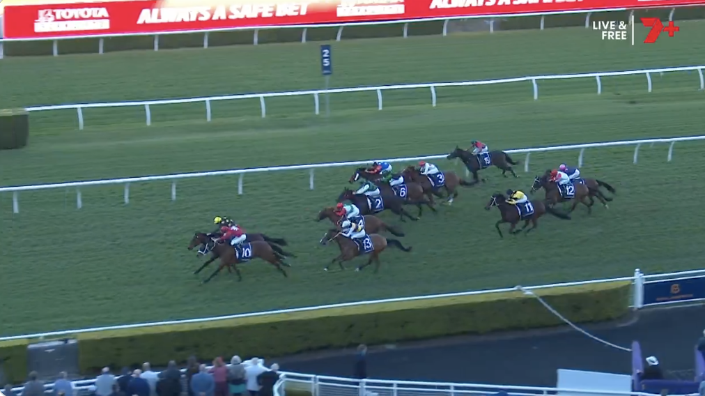 Horse race on a green turf track with jockeys in colorful silks chasing the lead near the inside rail; digital ad boards in background.
