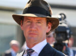 Man in a brown fedora, pinstripe suit, and tie at an outdoor event, facing the camera.