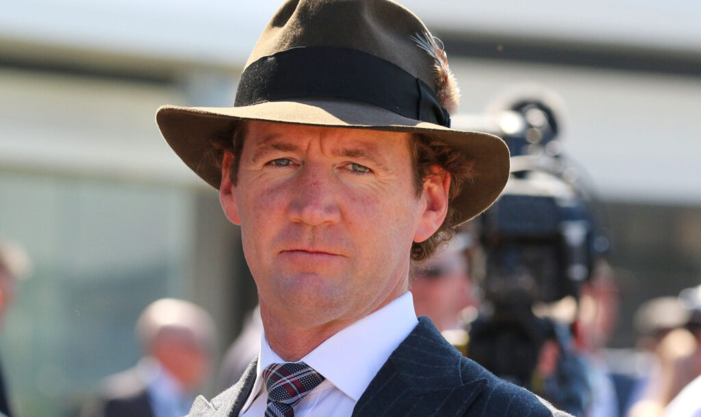 Man wearing a brown fedora and pinstripe suit at an outdoor formal event, with a camera crew out of focus in the background.