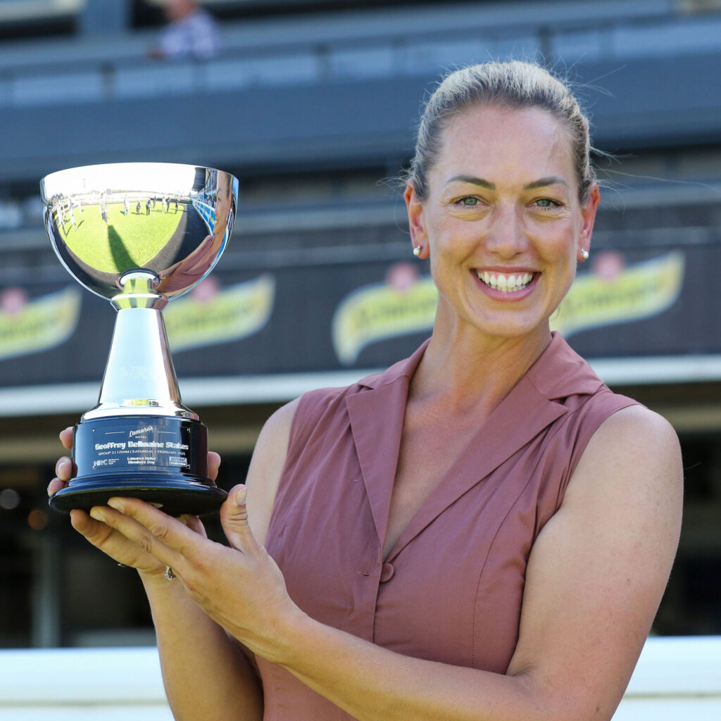 Smiling woman in a mauve blouse holding a shiny trophy outdoors.