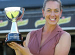 Smiling woman in a mauve blouse holding a shiny trophy outdoors.