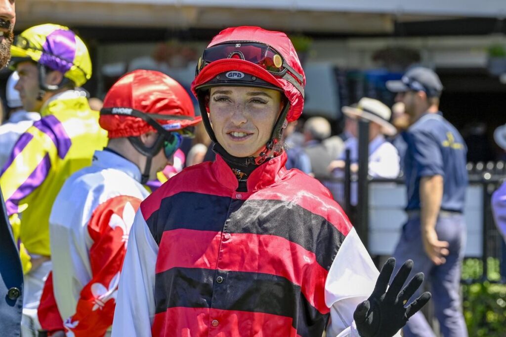 Person in red-and-black striped racing silks and red helmet waves from a paddock with others in colorful gear in the background, at a horse racing venue.