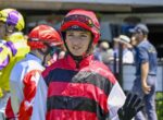 Person in red-and-black striped racing silks and red helmet waves from a paddock with others in colorful gear in the background, at a horse racing venue.