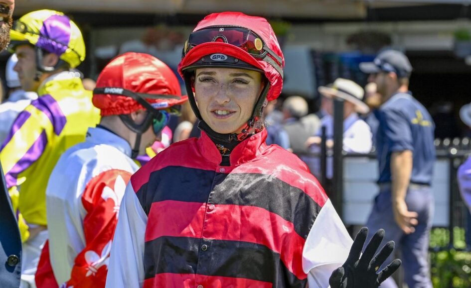 Person in red-and-black striped racing silks and red helmet waves from a paddock with others in colorful gear in the background, at a horse racing venue.
