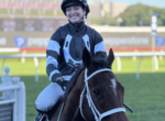 Jockey in black-and-blue silks and helmet smiles while riding a dark horse at a racetrack.