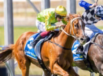 Two jockeys ride racehorses at full speed in a close horse race on a sunny track, leaning forward.