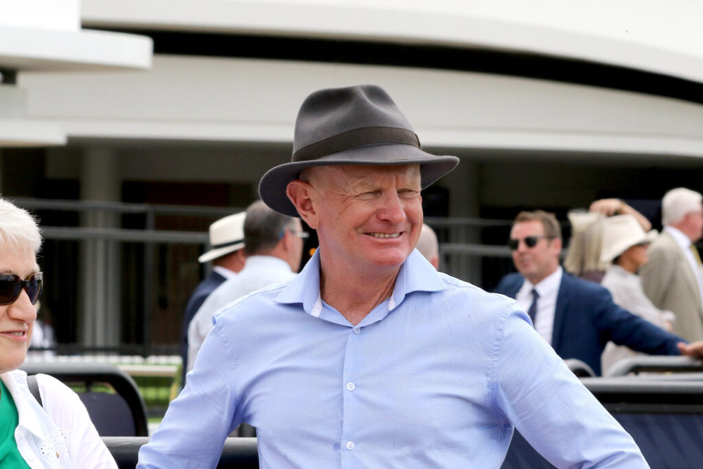 Smiling man in a light blue shirt wearing a gray fedora at an outdoor event, with others in the background.